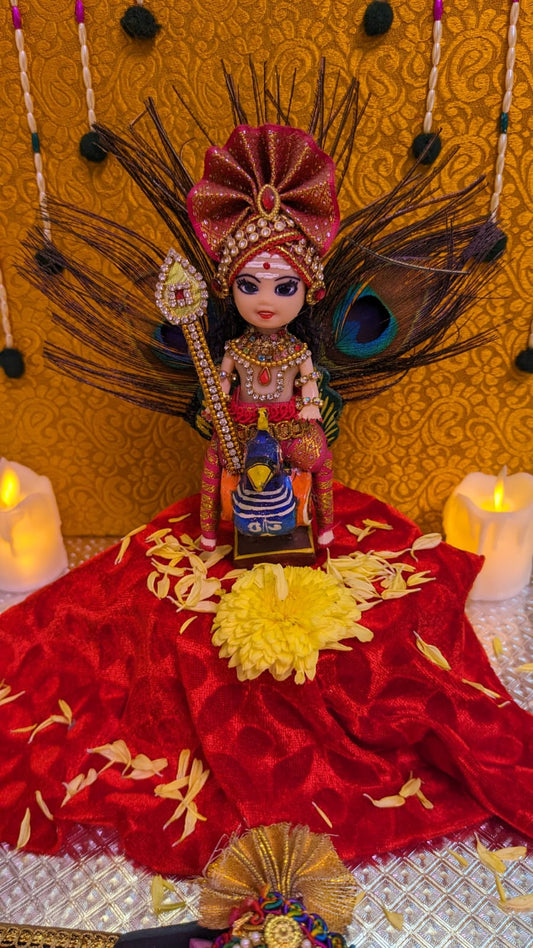 Handcrafted doll of Lord Murugar seated on a peacock, adorned with jewelry and dressed in red attire, displayed against a decorative background.